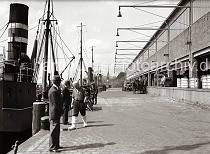 804_B_287 Rechts die 1937 neu gebaute Fischauktionshalle am Kai des Altonaer Fischereihafens -  Tonnen und Holzkisten zum Transport der Fische stehen auf der Laderampe des Gebudes. Ein Pferdefuhrwerk wird beladen, dahinter ein Zugmaschine mit Anhnger, die von Kaiarbeitern mit Kisten beladen wird. Mehrere Fischdampfer mit hohem Schornstein liegen am Kai - einer der Fischer geht mit heruntergeklappten Fischer-Gummistiefeln ber die Kaianlage. 