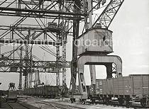 825_B_260a Am Kohlekai des Harburger Seehafens stehen offene Güterwagen unter dem Kohlekran und dem Portalkran. Ein Kohlefrachter liegt am Kai und die Kohleladung wird gelöscht - einer der Greifer schwebt gerade über einem der Güterwagen. Einer der Greifer steht abmontiert auf einem Bock bei den Eisenbahngleisen.