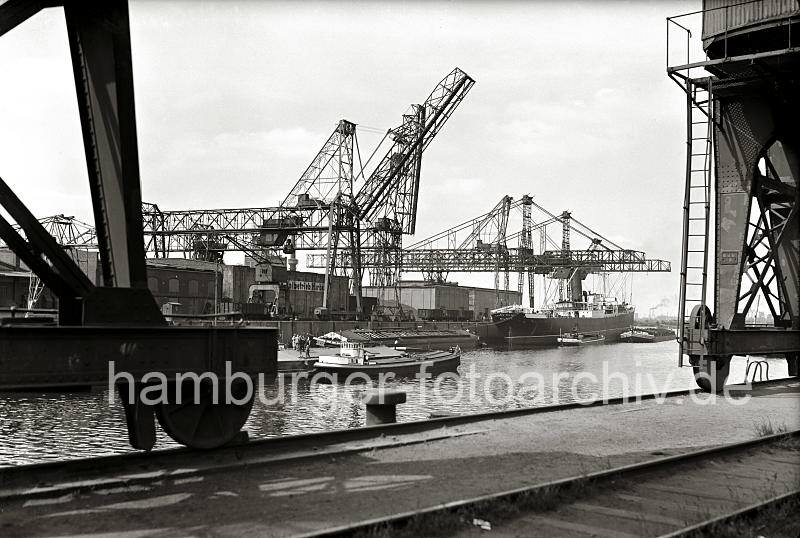 Alte Harburger Hafenaufnahmen - Krananlagen Seehafen Harburg - Binnenschiffe und Dampfschiff; ca. 1938 - Alte Harburg Bilder als Geschenk zum Geburtstag, Jubilaeum + Weihnachten. 828_B_262 Blick ber das Hafenbecken des Harburger Seehafens; im Vordergrund die Rder auf denen die Halbportalkrne auf der Kaimauer entlang gefhrt werden. Auf der gegenber liegenden Seite des Hafenbeckens hat eine Schute sowie ein Binnenschiff lngsseits eines Kohlenfrachters festgemacht. Ein mit Kohle gefllter Greifer schwebt ber dem Lastkahn - lks. ein Elbkahn, durch dessen geffneter Abdeckung die Ladung Kohle zu erkennen ist.
