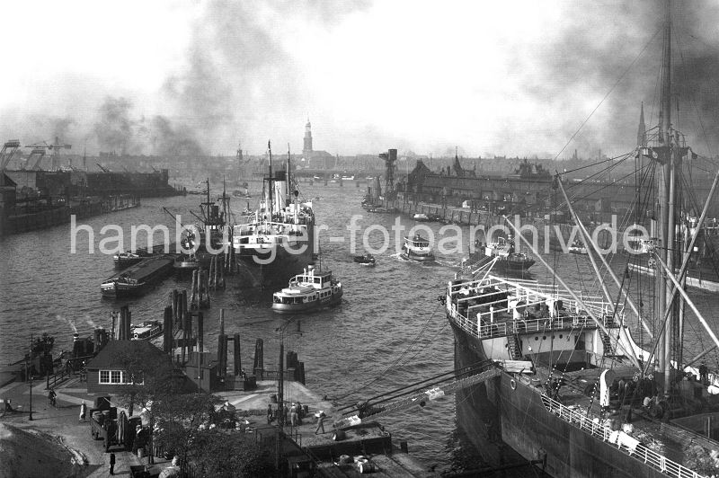 historische Hafenbilder aus dem Archiv der Hamburger Hafen und Logistik AG, HHLA.  176_130 Ein Frachter wird am Hansahft entladen - eine Gangway fhrt zum Kai, ein Arbeiter kommt von Frachtwagen und trgt Ware zum Schiff. Ein weiteres Frachtschiff liegt an den Dalben, eine Hafenfhre fhrt Richtung Elbe. Im Bildzentrum ragt der Turm vom Hamburger Wahrzeichen, der Michel zwischen den Huserdchern heraus. Links davon ist der Uhrturm vom Kaiserspeicher zu erkennen. Auf der rechten Bildseite die Einfahrt zum Hansahafen und Lagerschuppen am O'swaldkai, rechts der Indiahafen mit dem Afrikakai.