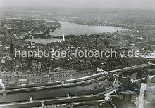 266_0431 Luftaufnahme ca. 1930; ganz im Vordergrund ist ein Ausschnitt vom Grasbrookhafen mit den Lagerschuppen des Dalmannkais zu erkennen.  Dahinter der Sandtorhafen mit den Schuppen am Sandtorkai, die sich bis zur Brooktorschleuse hinziehen. Dann folgt nach der Schlickschleuse der Brooktorhafen und der Kaispeicher B. Hinter der hohen Backsteinarchitektur der Speicherstadt liegt die Hamburger Altstadt mit vier Hamburger Hauptkirchen St. Nikolaikirche, St. Katharinen, St. Petrikirche und die St. Jakobikirche (v. lks.) In Nhe der Binnenalster das Hamburger Rathaus mit Turm - Segelschiffe fahren auf der Auenalster.