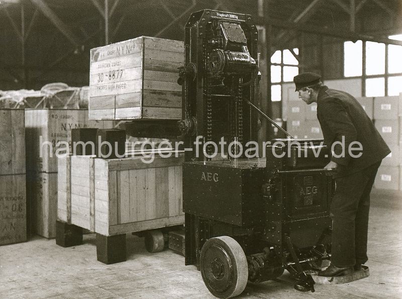 307_54 Der Lagerarbeiter stellt mit Hilfe eines Elektroschubbkarrens eine grosse Holzkiste in einem Lagerschuppen des Hamburger Hafens ab; rechteckig Holzblcke wurden als Abstandshalter zwischen den einzelnen Kisten verwendet. Die klobige Maschine erleichtert die schwere Arbeit der Hafenarbeiter und ist mit seiner Arbeits-Technik als Vorlufer des Gabelstaplers anzusehen. Im Hintergrund ist die Holzstnder-Konstruktion unter dem Dach des Hafenschuppens zu erkennen.