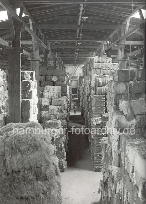 310_3987 Wollballen in einem Lagerschuppen im Hamburger Hafen - die Wolle ist zu Bndeln zusammen gebunden  fast bis an die Holzdecke der Lagerhalle gestapelt. Unter dem Dach angelegte Fenster sorgen fr die notwendige Durchlftung und Lichteinfall. Im Hintergrund steht ein Arbeiter im Durchgang zwischen den Wollballen.