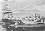 0953974 Blick vom Segelschiffkai in den Segelschiffhafen,  ca. 1890; links der Amerikakai und rechts der Asiakai. Khne haben lngsseits an den Frachtschiffen fest gemacht und werden mit der Fracht der Segelschiffe beladen. Diese Binnenschiffe transportieren ihre Fracht ber die Elbe in das Hinterland. Im Vordergrund rudern zwei Schiffer in einem hlzernen Ruderboot zu ihrem Kahn - um die Ladung trocken zu halten wird die Luke mit Brettern abgedeckt. 