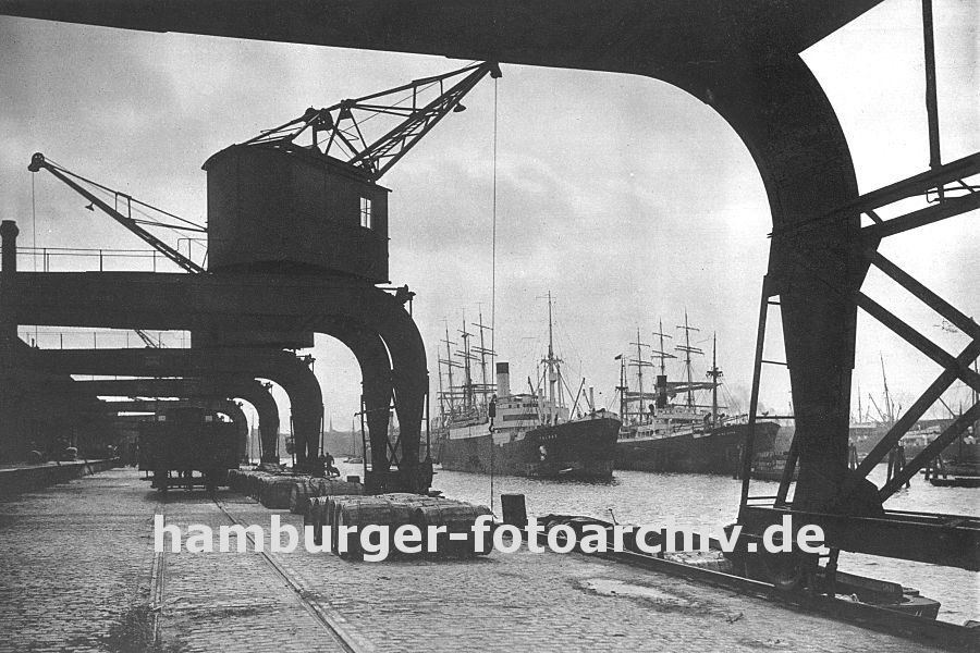 0953978 Krne am Amerikakai im Segelschiffhafen des Hamburger Hafens - ein Gterwagen der Hafenbahn steht auf den Gleisen am Kai und wird beladen. Fsser aus Holz liegen auf den Pflastersteinen und warten darauf in die am Kai liegenden Schuten verladen zu werden. Schiffe liegen auf Reede, fest gemacht an den Duckdalben aus Holzstmmen, im Hintergrund sind die Masten zweier Segelschiffe, Frachtsegler zu erkennen