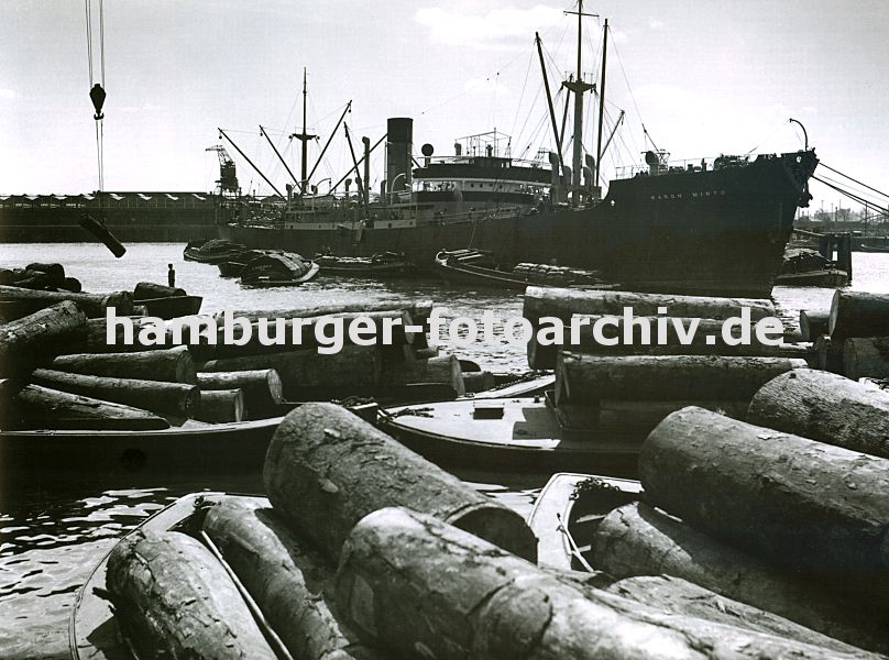 0953991 Ein Frachtschiff liegt im Sdwesthafen auf Reede; seine Ladung, die in Scken verpackt ist wird gelscht und mit bordeigenem Ladegeschirr auf Schuten verladen - im Hintergrund die Lagergebude am Windhukkai. Im Vordergrund werden am Togokai Schuten mit Baumstmmen beladen.