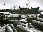 0953991 Ein Frachtschiff liegt im Sdwesthafen auf Reede; seine Ladung, die in Scken verpackt ist wird gelscht und mit bordeigenem Ladegeschirr auf Schuten verladen - im Hintergrund die Lagergebude am Windhukkai. Im Vordergrund werden am Togokai Schuten mit Baumstmmen beladen.  