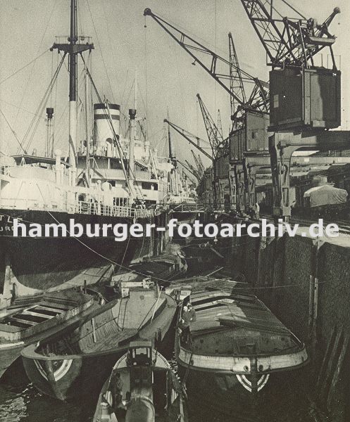 0953998 Frachter am Auguste Victoria Kai im Kaiser Wilhelm Hafen im Hamburger Hafen. Schuten liegen an der Kaimauer und knnen so mit der Fracht beladen werden. Im Vordergrund bewegt eine Barkasse einen leeren Frachtkahn. Auf dem Kai stehen Krne, die auf Schienen laufen und so die Ladung flexibel transportieren knnen - unter der Krananlage verlaufen die Schienen der Hafenbahn; die beladenen Gterwaggons bringen ihre Ladung in das Hamburger Hinterland.