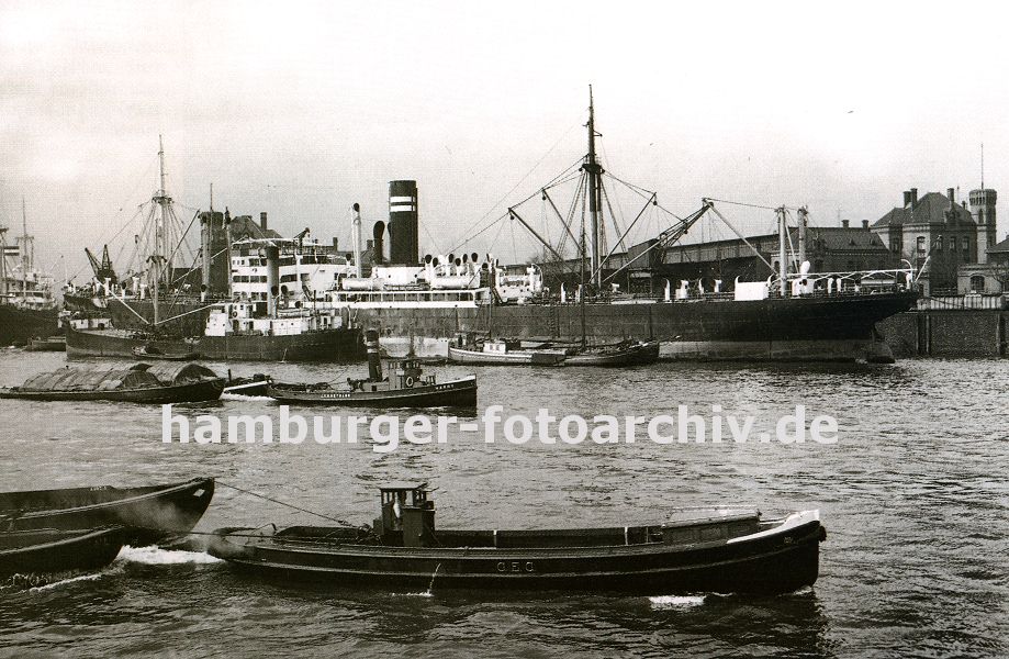 0954000 Schlepper ziehen jeweils zwei Schuten durch den Ellerholzhafen - ein Frachter liegt am Mnckebergkai und lscht seine Ladung auf lngsseits liegende Boote. Rechts im Hintergrund die im Hamburger Hafen typische Industriearchitektur mit Turm - das Gebude steht am Kronprinzenkai im Kaiser Wilhelm Hafen. 