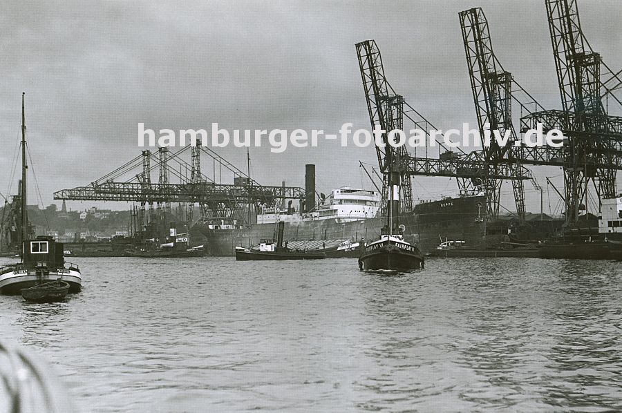 0954008 Im Hamburger Kohlehafen liegen die Kohlefrachter am Kai; die Ladung wird ber weit ausladende Krananlagen gelscht. iDie Ausleger der vorderen Krne sind hochgefahren - die Ladearbeiten sind beendet, Schlepper bereiten das Auslaufen des Schiffes vor. Links im Hintergrund Blick ber die Elbe nach Altona - der Kirchturm der Christianskirche ist zu erkennen. 