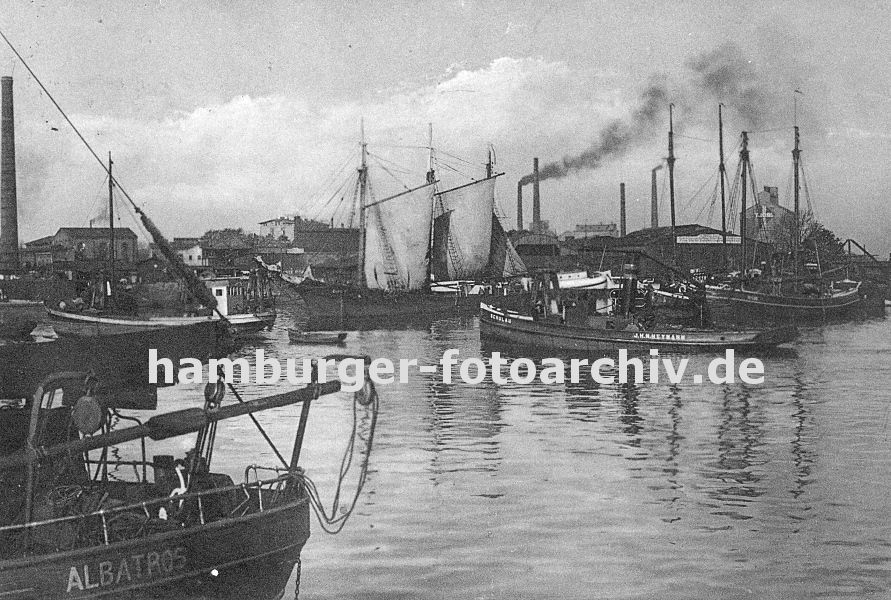 k_0954014 Blick in den Harburger Hafen ca. 1936 - der Dampf - Schlepper Schulau fhrt durch den Hafen Harburgs - Segelschiffe liegen am Kai. Ein Frachter hat Segel gesetzt und bereitet das Auslaufen vor - der Schlepper wird das Schiff von seinem Liegeplatz weg schleppen. Im Hintergrund qualmen hohe Schornsteine der Harburger Industrieanlagen.