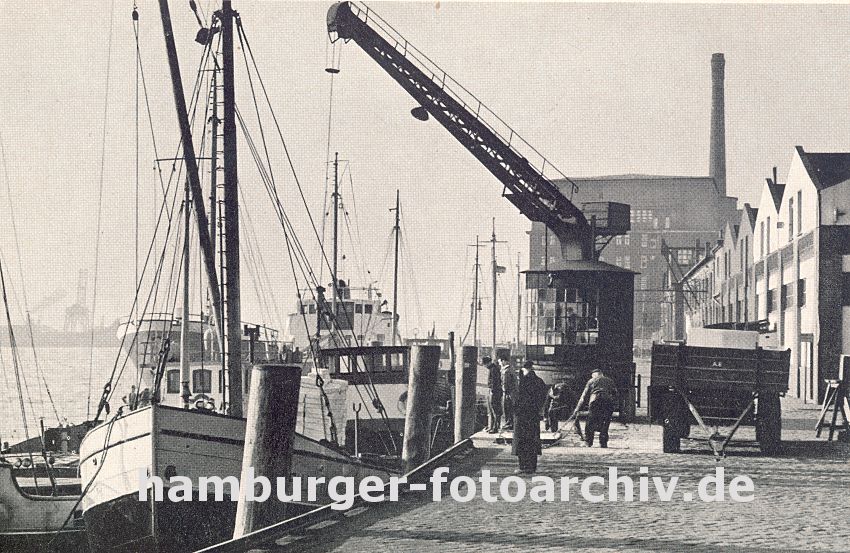 0954047 Bilder von Altona - historische Motive der Hafenstadt :: der Kai im Altonaer Hafen ist mit Kopfstein gepflastert - ein Kran holt die Ladung aus dem Laderaum des Frachtschiffs und ldt sie auf eine Anhnger der an Land steht; rechts Lagerhuser. einige Hafenarbeiter sehen der Arbeit zu,  andere bereiten dien Empfang der nchste Ladung vor. Lks. im Hintergrund auf der anderen Elbseite die Hafenanlagen von Waltershof.