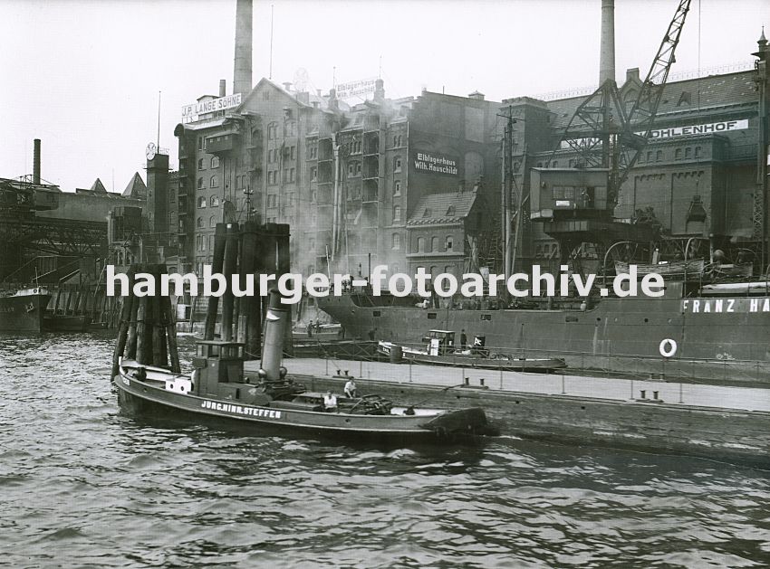 0954048 Altona Fotografie - Bilder der Stadt :: Speichergebude im Altonaer Hafen - ganz lks. eine Getreidemhle, ber lange Ladestutzen wird das Getreide von den Schiffen in das Gebude transportiert. Die Speichergebude sind in der fr die Zeit um 1900 typischen Industriearchitektur gehalten; in der Bildmitte ist das Elblagerhaus mit den Eingangsluken zu den einzelnen Lagerbden - die einzelnen Lagerbden mit den Eingangsluken werden mit Winden / Krnen die unter dem Gebudedach montiert sind,  beliefert. Auf der rechten Bildseite der Kohlehof / Kohlespeicher; der Greifer eines Krahns hat gerade eine Ladung Kohle vom Schiff geholt - aus dem Greifer rieselt links und rechts die Kohle heraus. Im Vordergrund liegt ein Schlepper am Ponton unter Dampf, dahinter Duckdalben, die aus Baumstmmen gefertigt und in den Hafenboden gerammt sind - an ihnen knnen Schiffe festmachen. 