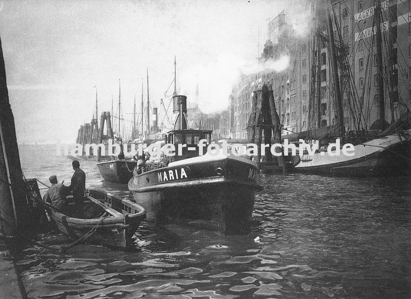 0954049 Hafenanlage des Altonaer Hafens - hohe Speichergebude und Lagerhuser stehen an der Wasserseite von der aus direkt von den Schiffen die Ladung gelscht werden kann. An einer langen Reihe von Duckdalben aus Baumstmmen / Tannenstmmen haben Schiffe festgemacht. Im Vordergrund re. ein Ruderboot aus Holz, die Ruder liegen im Boot. Zwei Hafenarbeiter blicken zum heran fahrenden Schlepper, der eine mit Scken beladene Schute zieht. 