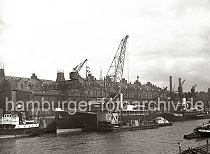 01181_11925T Ein Schwimmkran beldt den Frachter MARYLEBONE, der am Sandtorkai mit einer groen Holzkiste. Weiter Fracht liegt im Laderaum einer Schute zum Verladen bereit. Hinter den Kaischuppen sind die Dcher der Speicherstadt zu erkennen, auf der linken Seite die beiden Schornsteine vom Kesselhaus.