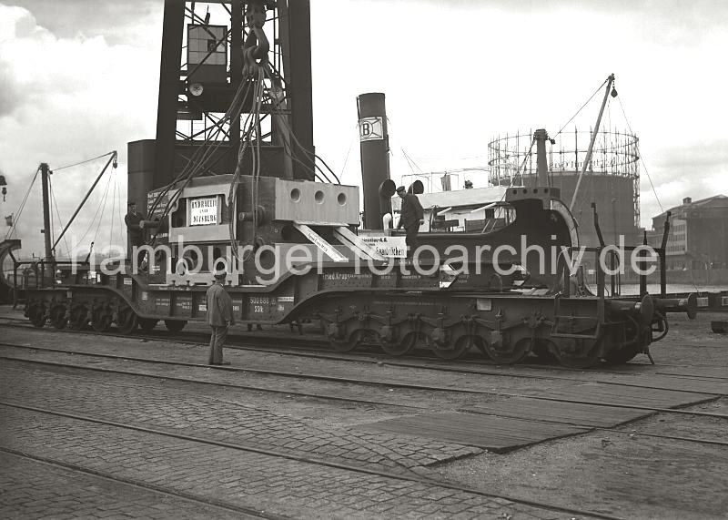 Historische Fotos aus dem Bildarchiv der HHLA; Verladung von Maschinenteilen im Hafen Hamburg - FotoPrints auf Leinwand, Posterprint als Geschenk fuers Jubilaeum.  02208_559 Ein Schwertransport-Gterwagen steht mit einem schweren Maschinenteil auf den Gleisen der Hafenbahn vor dem 150 Tonnen Kran am Kranhft. Kaiarbeiter sind dabei Eisenseile zum Heben der Last vorzubereiten. Auf der anderen Seite der Elbe Industrieanlagen der Gaswerke auf dem Grasbrook.