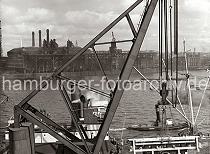 02208_559 Der groe Kran hebt ein Maschinenteil von der Kai- flche am Kranhft, der uersten Spitze des Segelschiffhafens. Der Dampfer, der die schwere Fracht an Bord nehmen soll, liegt an der Kaimauer - ein Schlepper, aus dessen Schornstein schwarzer Qualm steigt fhrt Richtung Veddelhft. Am gegenber liegenden Ufer der Norderelbe steht die Industriearchitektur der Gasanstalt und rechts ist die Einfahrt zum Magdeburger- und Baakenhafen zu erkennen.