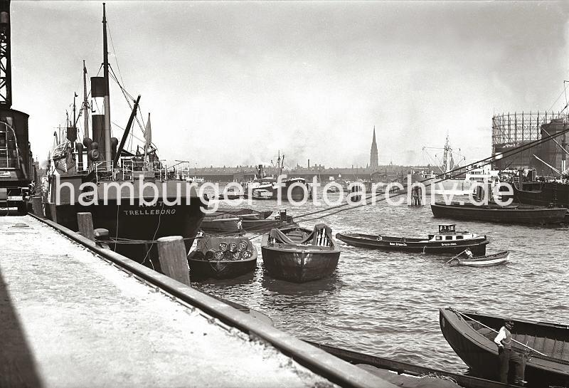 Historische Hafenmotive von der Hansestadt Hamburg; Schuten, im Laderaum Kantholz; Segelschiffhafen / Kaischuppen am Strandkai ca. 1935 - Fotos auf Foto-Leinwand, Posterprint als Geschenk fuer Jubilaeum, Abschied + Geburtstag.  02214_B_227a Am Amerikakai des Segelschiffhafens liegt der schwedische Frachter KNUT aus Trelleborg - die Ladung der Schuten ist teilweise mit Persenning gegen Witterungseinflsse abgedeckt. Ein Jollenfhrer wriggt seine Jolle durch das Hafenbecken. Im Vordergrund steht ein Ewerfhrer bei seiner Schute - den langen Peekhaken / Bootshaken, mit dem der Schiffsfhrer den antriebslosen Lastkahn stakend durch das Hafengebiet fhrt, hat er am Bug abgelegt. Im Hintergrund der Gasometer auf dem Grasbrook und die Schuppen am Strandhafen.