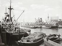 02215_604 Frachtschiffe liegen im Segelschiffhafen - der Gterumschlag findet zur Kaiseite mit Krnen statt; zur Wasserseite werden die Waren mit bordeigenen Ladebumen auf Schuten und Elbkhne geladen oder aufgenommen.  Die Ladungen der einfachen Schuten sind mit Planen abgedeckt, bei dem Lastkahn rechts im Vordergrund kann der Laderaum mit Brettern abgedeckt und so die Ladung geschtzt werden. Im Hintergrund re. das Fhrerhaus des neuen Krans am Kranhft und lks. neben dem Gasometer der Kirchturm der St. Petrikirche, die Jakobikirche und der Hamburger Michel.