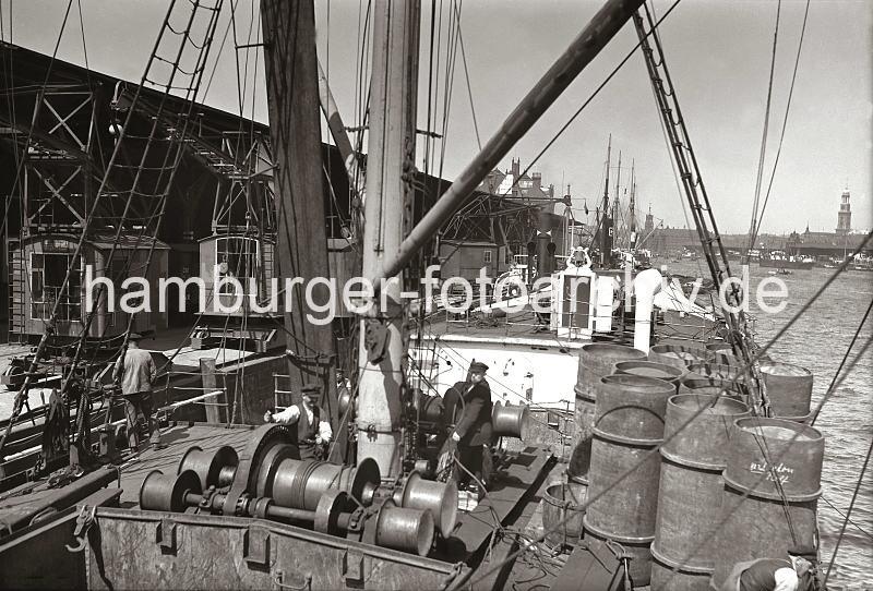 Historische Hafen Fotos aus der Hansestadt Hamburg; Deck eines Frachtschiffs im Segelschiffhafen - Rollkrne am Amerikakai - Fotos auf Foto-Leinwand, Posterprint als Geschenk fuer Jubilaeum, Abschied + Geburtstag.  02217_B_225a Blick ber das Deck eines Frachtschiffs im Segelschiffhafen - ein Arbeiter steht an der elektrischen Winde, die den Ladebaum des Schiffs betreibt. Im Hintergrund der hohe Uhrenturm des Kaispeichers A und hinter den Dchern der Kaischuppen des Strandhafens der Turm der St. Michaeliskirche.