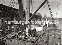 02217_B_225a Blick ber das Deck eines Frachtschiffs im Segelschiffhafen - ein Arbeiter steht an der elektrischen Winde, die den Ladebaum des Schiffs betreibt. Im Hintergrund der hohe Uhrenturm des Kaispeichers A und hinter den Dchern der Kaischuppen des Strandhafens der Turm der St. Michaeliskirche.