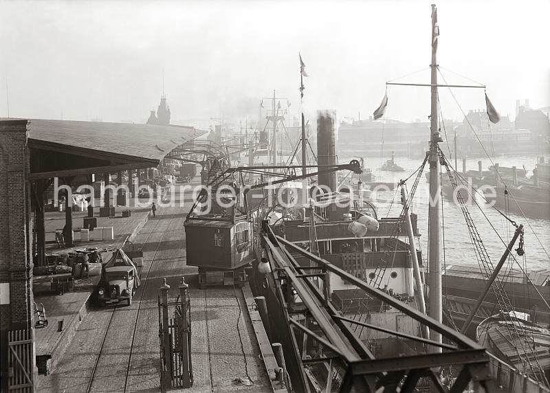 Historische Hamburgbilder aus dem Archiv der HHLA. Hafenbetrieb am Versmannkai des Baakenhafens - Warenumschlag am Hafenkai; ca. 1935. Alte Hamburg Fotos als Geschenk fr Bro und Wohnung; Fotoleinwand + Posterprints. 02228_793a Blick auf hinteren Abschnitt des Baakenhafen mit dem Schuppen 25 am Versmannkai. Ein Rollkran beldt einen Frachter mit Ballen - Kaiarbeiter bereiten auf der Laderampe den Transport weiterer Gter vor. In dem offenen Kaischuppen sind Kisten, Tonnen und Scke gelagert - der Zugang wird nachts mit einem Eisentor versperrt. Im Hintergrund sind in der Bildmitte und rechts die Bgen der Elbbrcken zu erkennnen und auf der linken Bildseite der niedrige Zinnenturm des Schleusenwrterhauses der Baakenschleuse, die zum Oberhafen / Billehafen fhrt.
