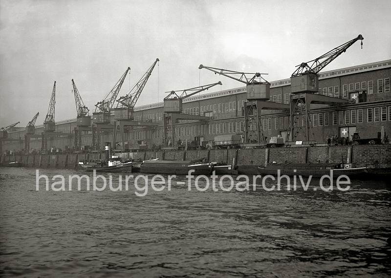 Historische Hamburgbilder aus dem Archiv der HHLA. Hafenschuppen und Halbportalkrne am Versmannkai / Baakenhafen; ca. 1935. Alte Hamburgbilder als Geschenk zur Erinnerung fr Bro und Wohnung; Fotoleinwand + Posterprints. 02229_189 Schuten liegen am Versmannkai des Baakenhafens an der Kaimauer vor dem Lagerschuppen 24. Die Laderume der Lastkhne sind gegen Witterungseinflsse abgedeckt. An der Laderampe werden Lastwagen und Pferdefuhrwerke mit Fruchtkisten beladen.