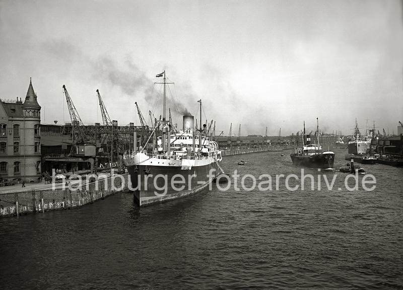 02236_152a Blick in den Baakenhafen; links der Versmannkai mit den Fruchtschuppen und dem Backsteingebude und dem Giebeltrmchen; der Frachtdampfer NATIA liegt am Kai. Rechts der Petersenkai mit Schiffen vor den Kaischuppen. Ein Kstenfrachter wird von einem Schlepper zur Elbe geschleppt, direkt daneben ein kleine Jolle, die vom Jollenfhrer durchs Hafenbecken gerudert wird. Historische Hamburger Hafenbilder. Fruchtschuppen mit Frachter am Versmannkai - Dampfschiffe am Petersenkai; ca. 1935. 