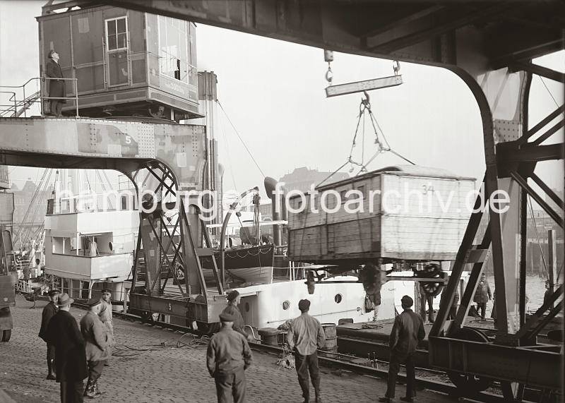 Hamburg-Hafenarchiv mit Fotos der HHLA / Hamburger Fotoarchiv Christoph Bellin. Zirkuswagen im Magedeburger Hafen - Frachter lscht Ladung; ca. 1935.  02253_579 Die Ladung eines Frachter wird vor dem Kaischuppen C des Magdeburger Hafens gelscht. Ein Zirkuswagen hngt an dem Haken eines Hafenkrans und wird an Land gebracht. Kaiarbeiter stehen mit den Hnden in den Taschen un der dem Kranportal und sehen der Arbeit zu.