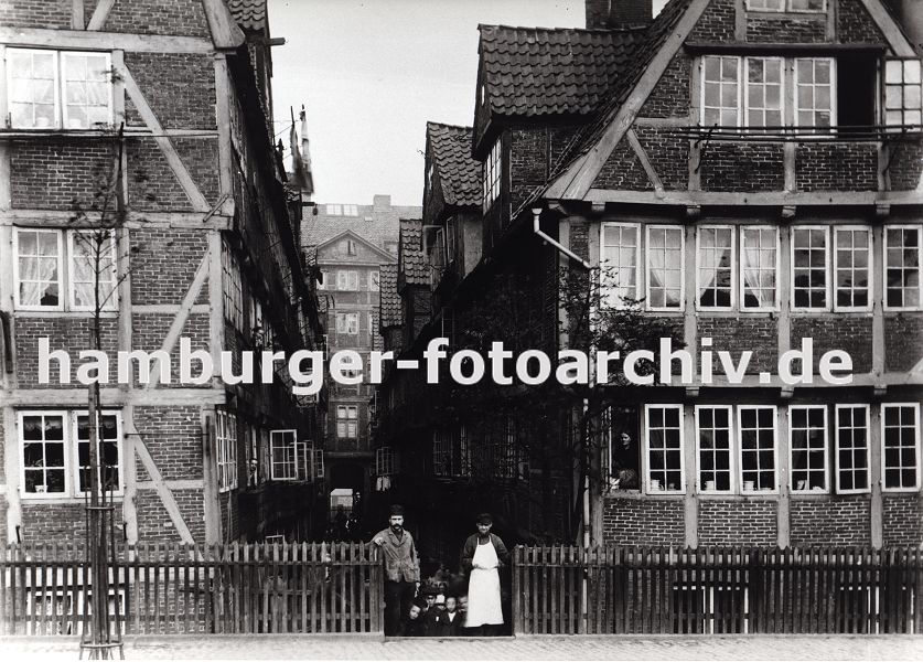 01147710 Hamburgensie, Hamburgs Vergangenheit - Bilder/Fotos :: Blick in einen Hof im Bereich Kehrwieder - Fassaden von Fachwerkhusern. Ein kleiner Strassenbaum ist mit einem Metallgitter geschtzt, in dem Tor von einem Holzzaun stehen zwei Handwerker / Arbeiter; einer von ihnen trgt eine weisse Schrze, im Hintergrund drngen sich neugierige Kinder. Die alten, schiefen Huser stehen dricht gedrngt, Wsche hngt zum Trocknen an einem Gestell aus dem Fenster. Durch den Zollanschluss Hamburgs 1888 an Preuen entstand der Hamburger Freihafen - ca. 20 000 Menschen, die auf den Elbinseln Kehrwieder, Brook und  Wandrahm lebten wurden zwangsumgesiedelt, die Wohn- und Lagerhuser wurden fr den Bau der Speicherstadt abgerissen. 