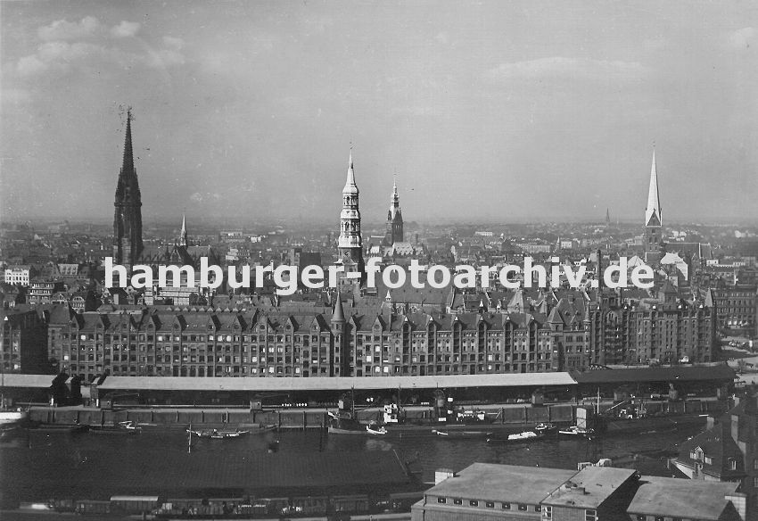 01147721 Bilder von Hamburg :: Blick ber den Sandtorhafen und dem Sandtorkai zur Hamburger Speicherstadt mit ihren Giebeln und Trmen der Backsteinarchitektur. Schiffe liegen am Kai und werden entladen und die Ladung in den Lagerschuppen am Kaispeicher verstaut. Hinter den Speichern das Panorama Hamburgs mit seinen markanten Trmen: links die Nikolaikirche, dann folgt der Turm der Hauptkirche St. Katharinen, deren Kirchspiel auch fr die Speicherstadt zustndig ist. Rechts daneben der Rathausturm und am rechten Rand der Turm der Hamburger St. Petrikirche.