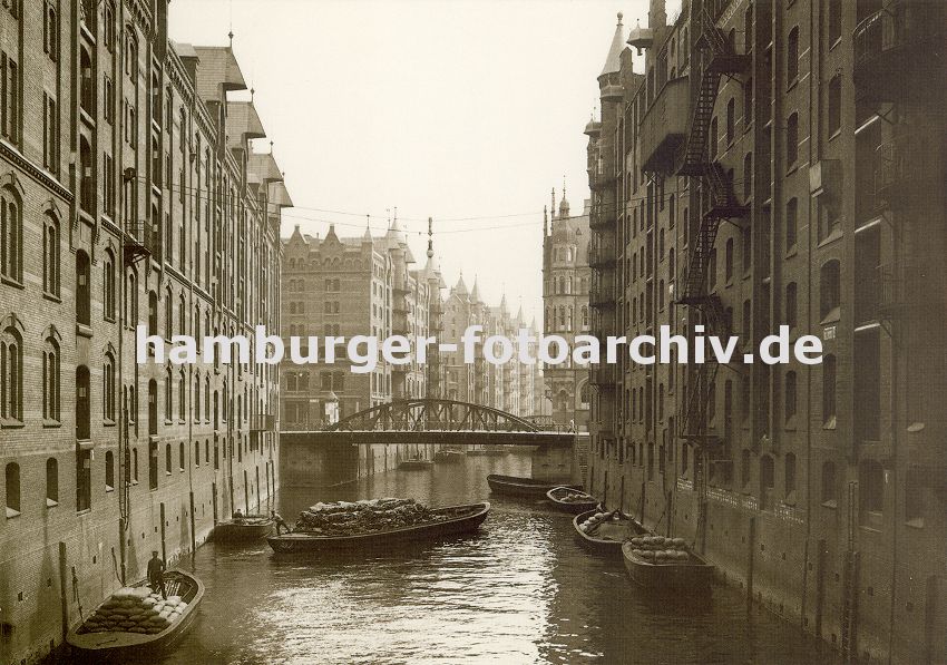 01147736 Schuten im Fleet in der Hamburger Speicherstadt - die Khne sind mit Scken beladen und sind an den Ringen, die in der Speichermauern eingelassen sind fest gemacht. Eine ber dem Wasser hngende Lampe soll die Arbeiten in der Dunkelheit erleichtern. Neben dem eisernen Rundbogen der Brcke ber den Fleet steht eine Litfasule. 