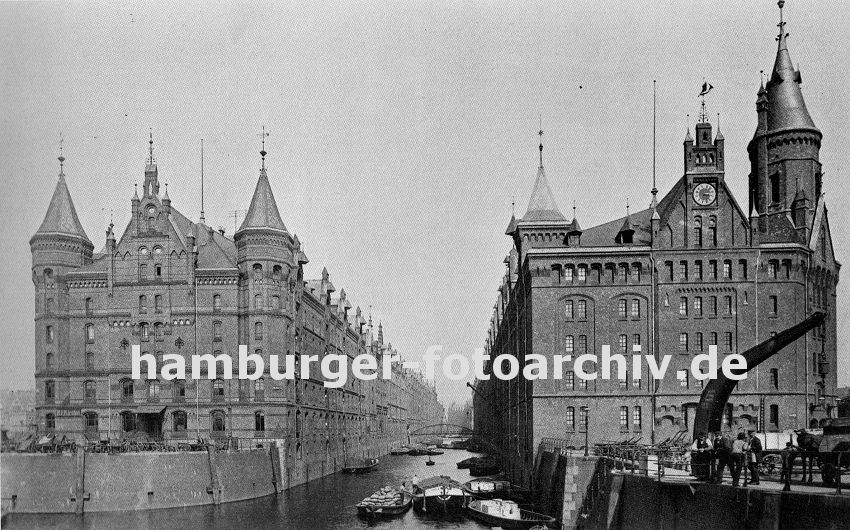 01147741 Hamburgensie - historische Fotografie - Blick auf die Speicherblocks A und J  an der Kehrwiederspitze; rechts ein stationrer Kran, dahinter sind Handkarren und Pferdewagen abgestellt, die fr den Transport von Waren innerhalb der Speicherstadt verwendet werden. Oben am Giebel ist des rechten Speichers unter dem Eisendekor eines Segelschiffes eine grosse Uhr angebracht. Auf dem Kehrwiederfleet liegt eine Kastenschute, ein Kahn hat lngsseits festgemacht und lscht seine Ladung mit Scken.