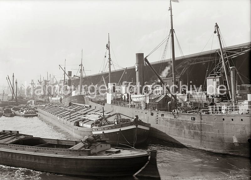 Hamburger Hafenfotografien aus dem Bildarchiv der Hamburger Hafen und Logistik AG HHLA. Frachtschiffe und Binnenschiffe am Dalmannkai / Grasbrookhafen; ca. 1936. 603_13_680a Gterumschlag am Dalmannkai des Grasbrookhafens; die Frachtschiffe liegen dicht an dicht an der Kaimauer und werden ber die Hafenkrane be- und entladen. In der Bildmitte ist das Motorschiff HOLSTENAU zu erkennen. Daneben liegen mit Kisten und Fssern beladene Schuten.  Im Vordergrund Binnenschiffe, die z.B. im Schleppverband die Gter auf der Elbe transportieren.